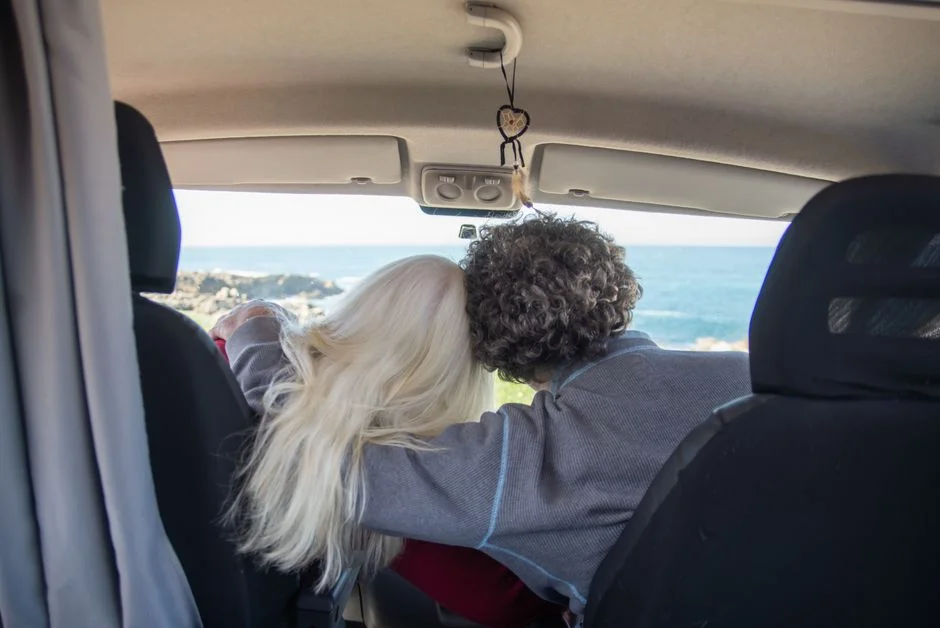 Paar sitzt im Innenraum eines Campervans und blickt durch die verglaste Heckklappe nach draußen aufs Meer