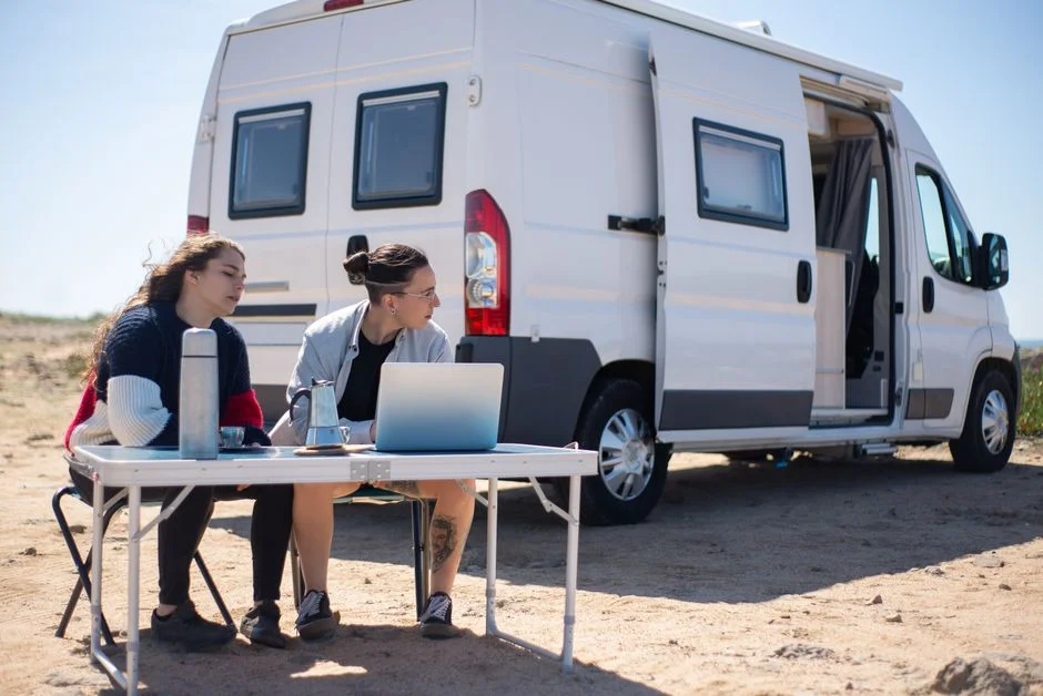 Zwei Personen arbeiten am Campingtisch mit Laptop vor einem weißen Kastenwagen